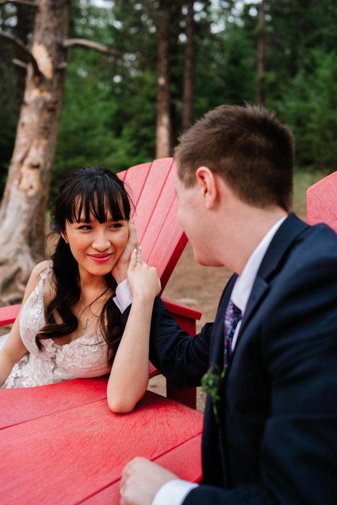 Bride looks at groom on red chairs in Jasper National Park