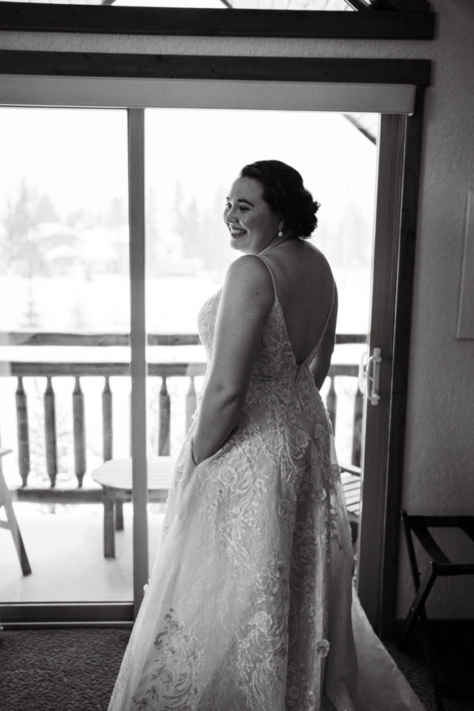 A bride stands infront of the window in Canmore Alberta with her hands in her dress pockets. 
