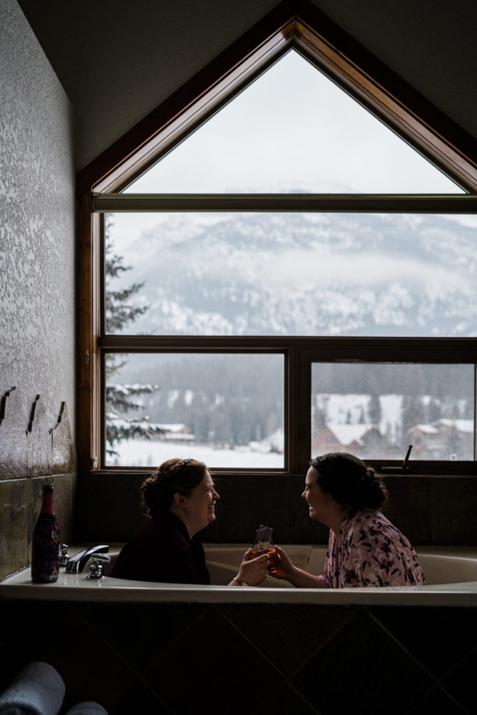 Two brides sit in a bathtub drinking wine on the morning of their wedding.
