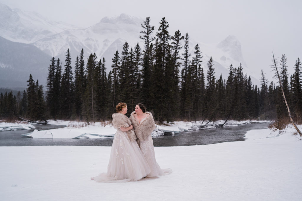 Two brides stand outside near the river in Canmore Alberta. 