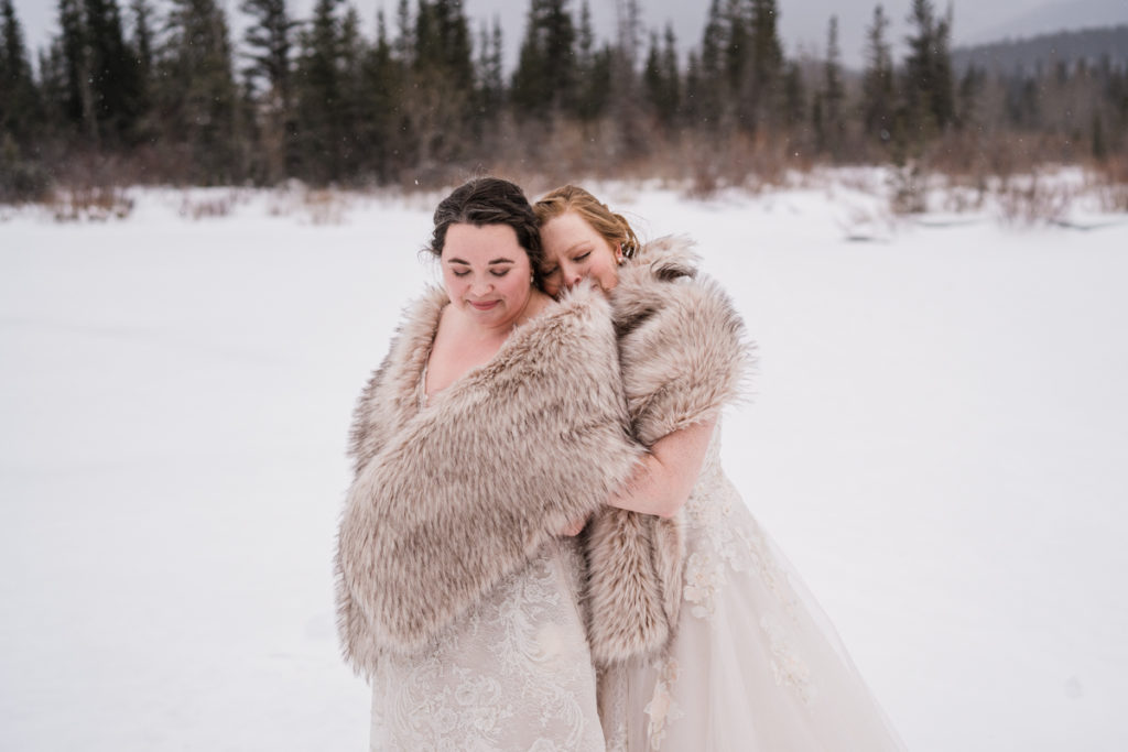 Two brides wrapped in fur shawls hug during winter wedding in Alberta. 