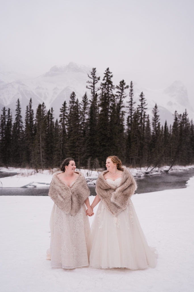 Two brides hold hands by river in Canmore Alberta in winter. 