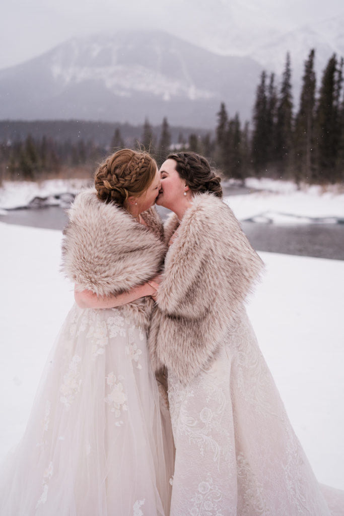 Two brides embrace while wearing fur shawls for winter wedding in Canmore.