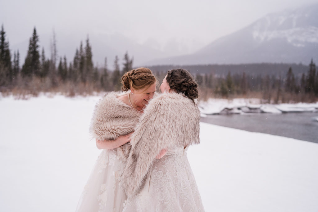 Two brides laugh together in front of Three Sisters mountains in Alberta.