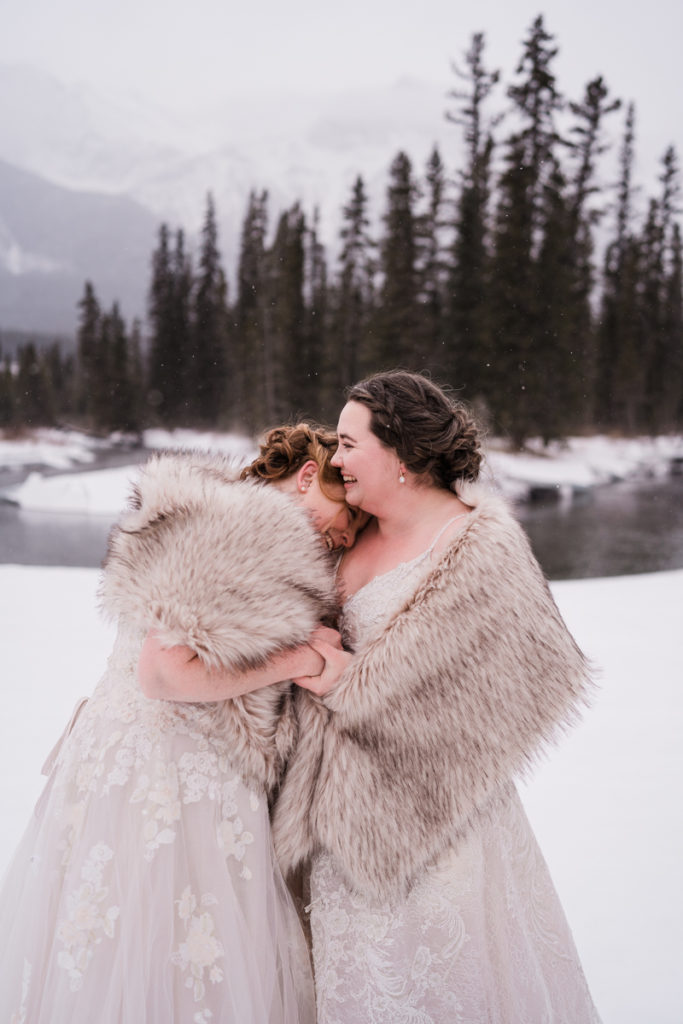 Two brides laugh during snowy winter wedding in Alberta. 