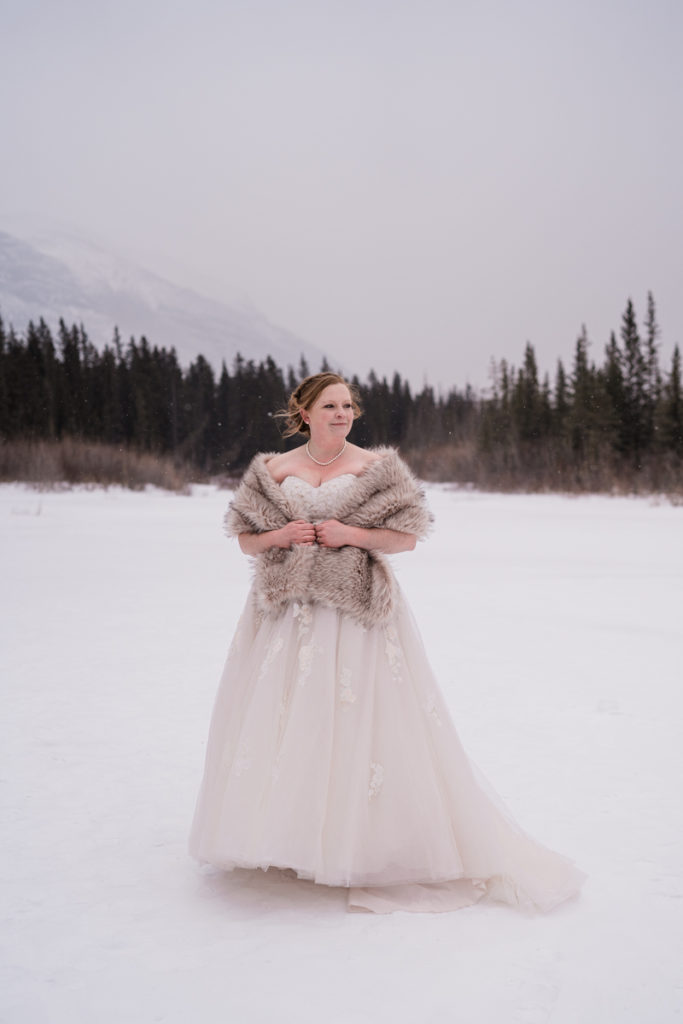 Bride wrapped in fur shawl looks off to the snowy mountains. 