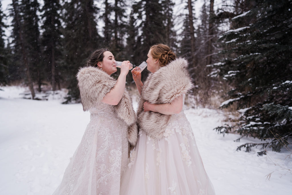 Two brides drink frozen champagne outside while wearing fur shawls. 