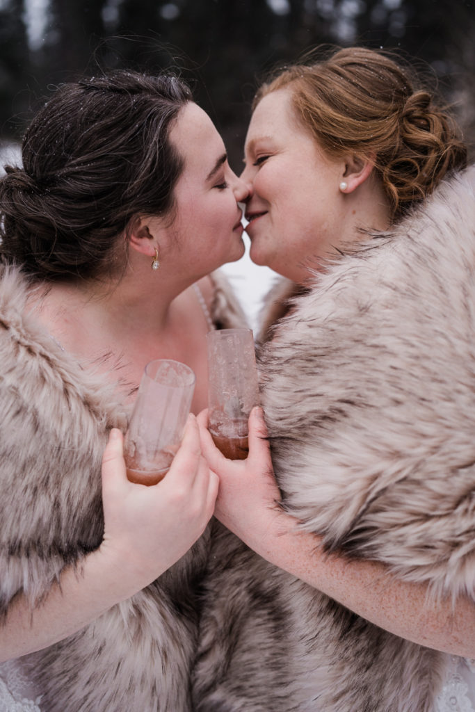 Two brides kiss in the snow while holding wine glasses.