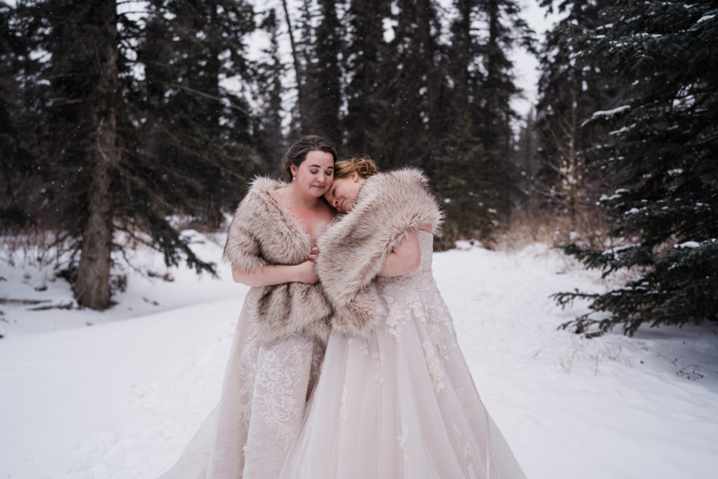 Two brides wearing fur shawls cuddle in the snowy trees in Alberta. 