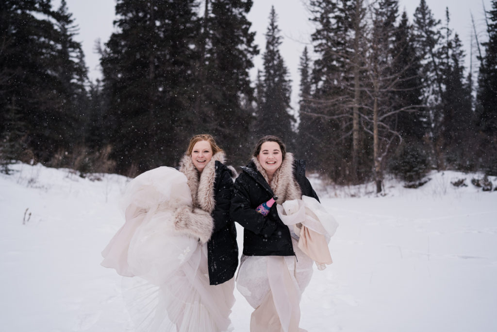 Two brides walk together while holding up their dresses during winter wedding in Canmore. 