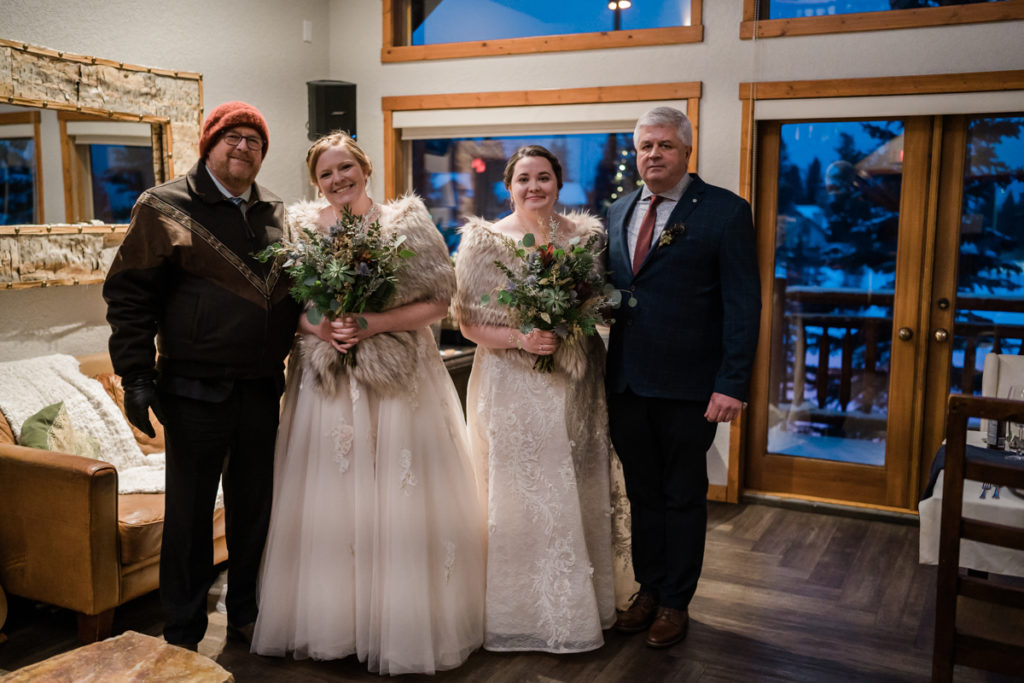 Two brides stand with their fathers before winter wedding ceremony. 