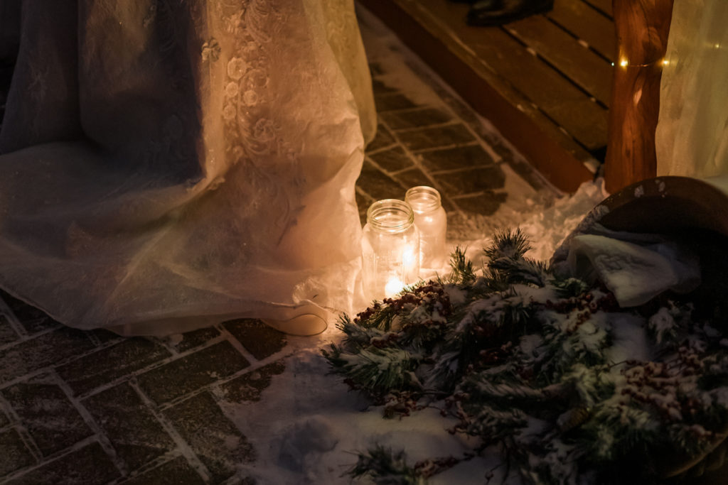 Wedding dress lays next to candles in mason jar and greenery in the snow