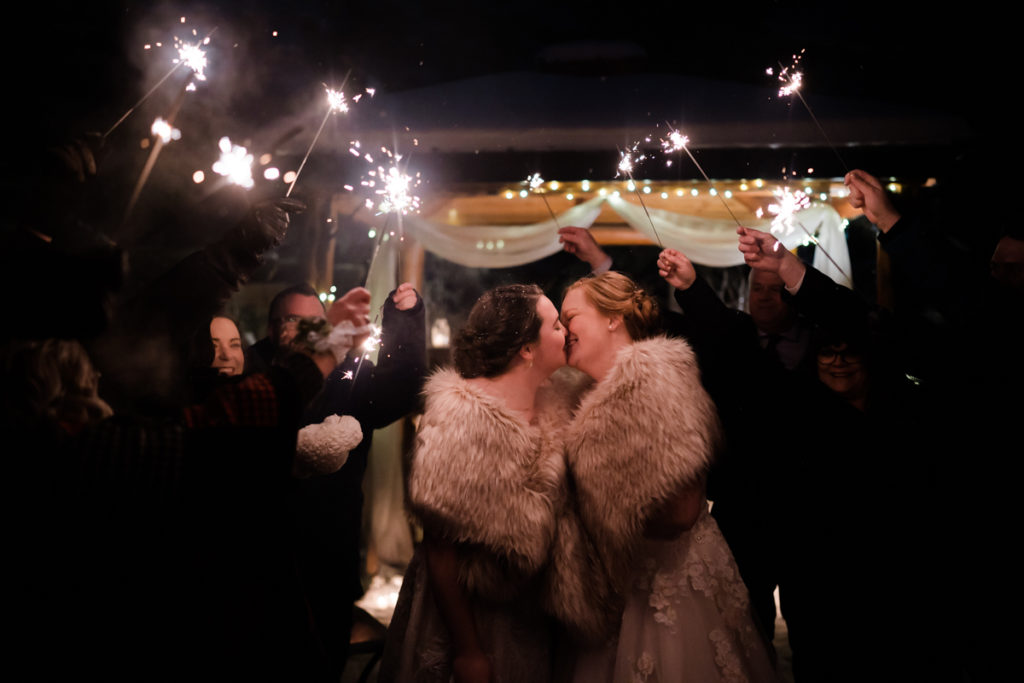 Two brides kiss during sparkler exit at outdoor winter wedding in Canmore
