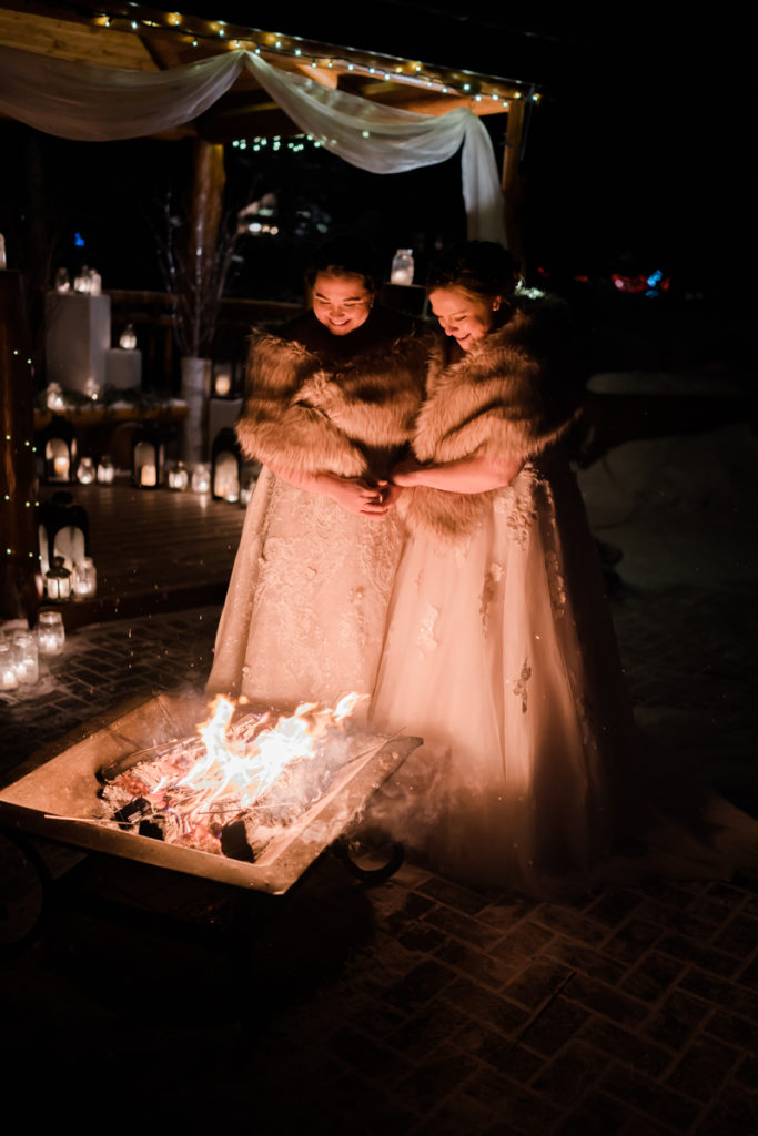 Two brides warm up infront of campfire after sunset winter ceremony. 