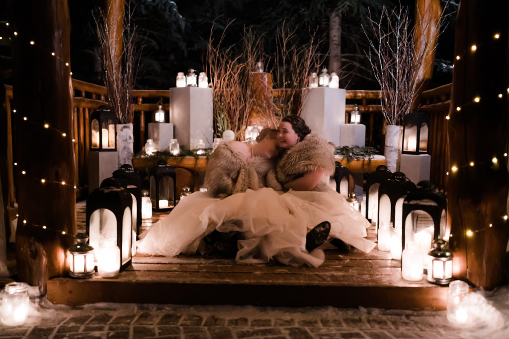 Two brides cuddle up in gazebo surrounded by candles and lanterns after sunset winter wedding. 