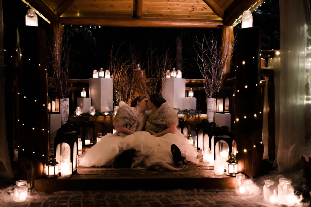 Brides wrapped in fur shawls kiss in candlelit gazebo after outdoor winter wedding