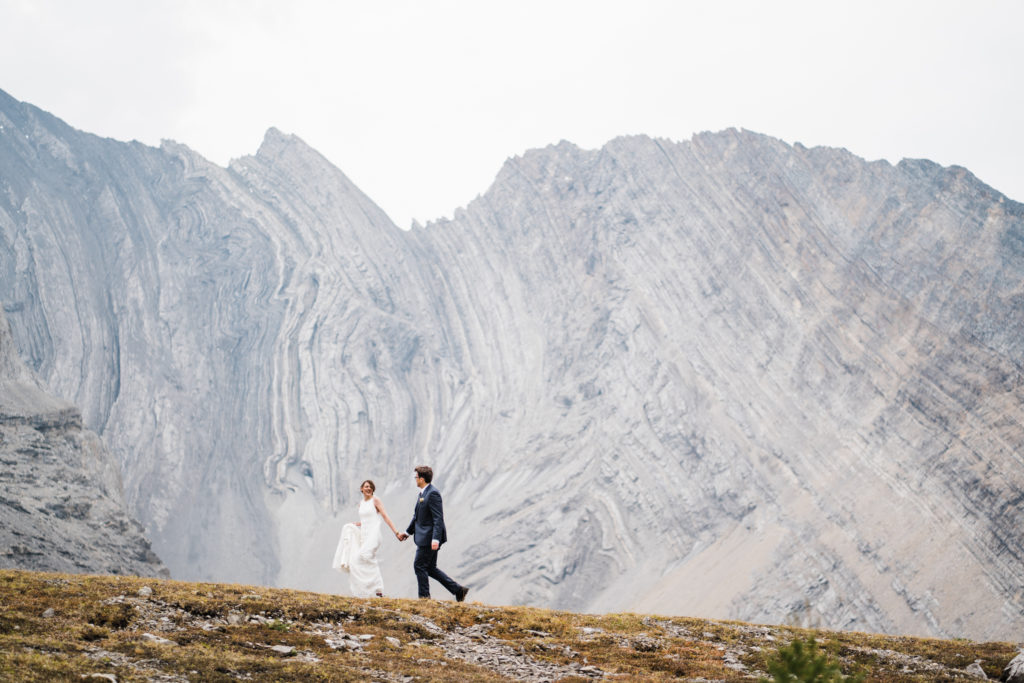 Bride leads groom walking along ridge with mountains in the background during their hiking elopement in Kananaskis. 