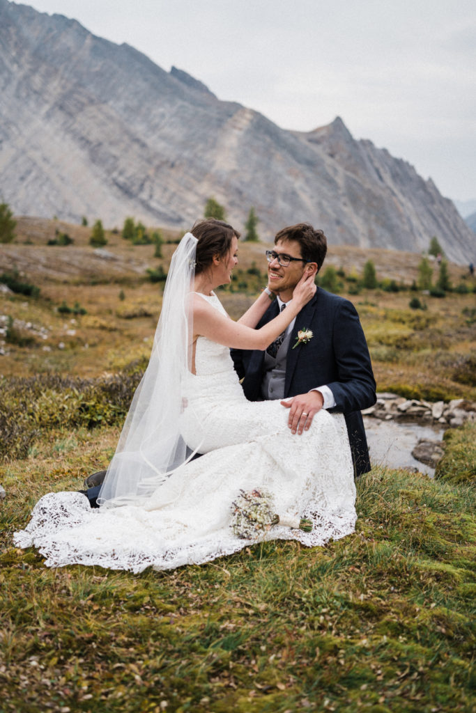 bride sits on groom lap in meadow in the mountains while smiling at each other. 