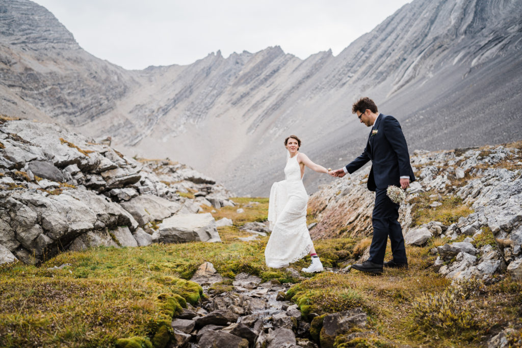 Bride leads groom over small river with towering mountains in the background.