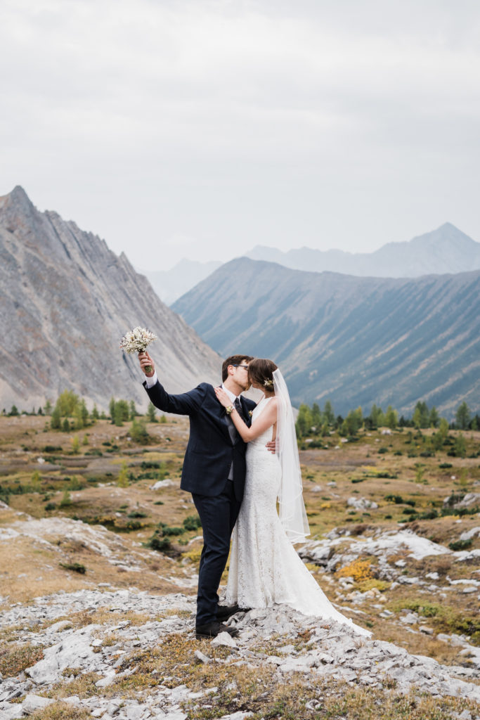 Bride and groom kiss while groom holds up bouquet during a hiking elopement in Kananaskis. 