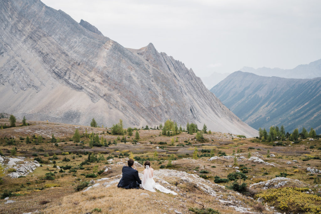 Bride and groom sit together in sub alpine meadow during hiking elopement in Kananaskis. 