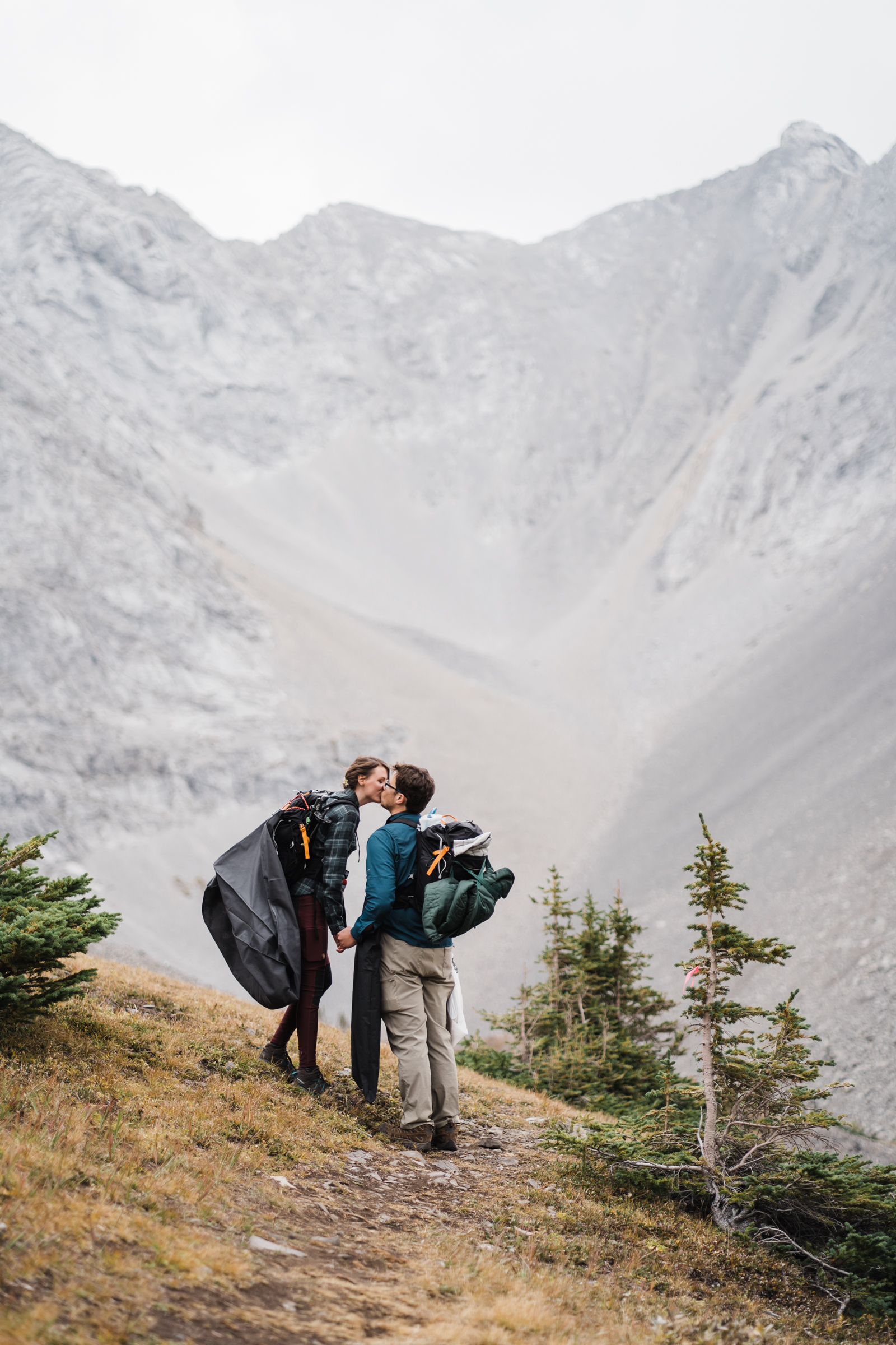 bride and groom carrying their wedding gear in their backpacks kiss infront of towering mountains. 