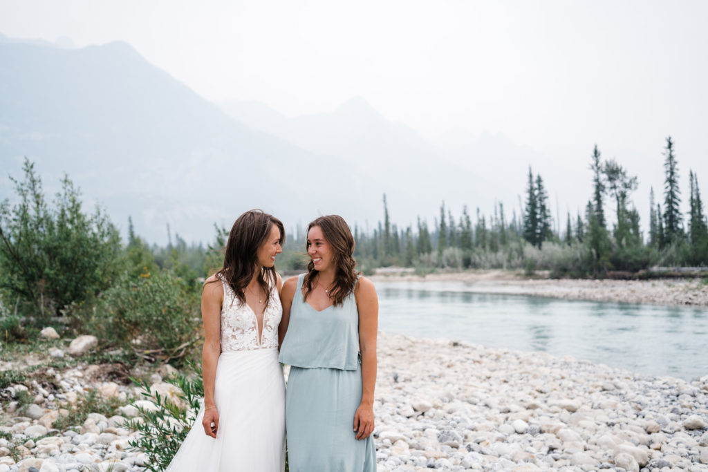 Bride and sister look at each other smiling in front of the Snaring river in Jasper National Park. 