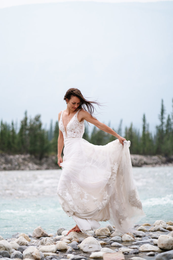 Barefoot bride holds her lace wedding dress up by river in Jasper National Park. 