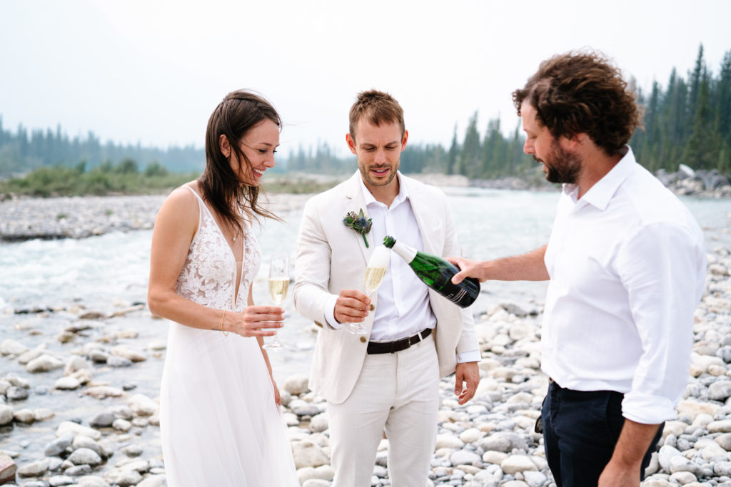 Man pours bride and groom champagne in front of Snaring River in Jasper National Park. 