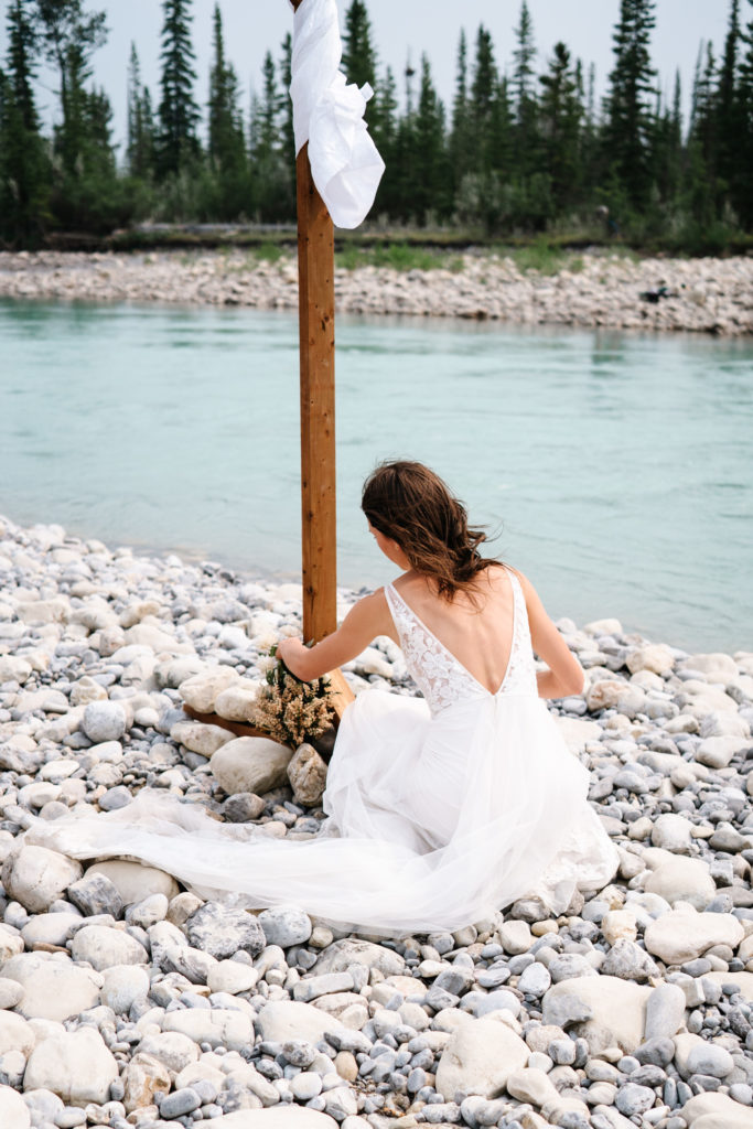 Bride places flowers next to wood wedding arch by Snaring River in Jasper National Park.