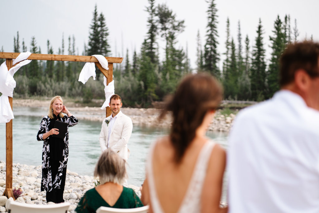 Groom looks at bride walking down towards him during intimate wedding ceremony in Jasper National Park. 