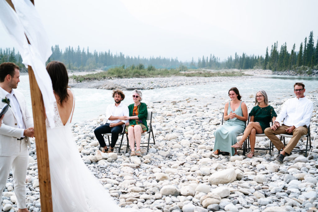 Family sits on chairs on rocks next to Snaring River during Jasper Intimate Wedding. 