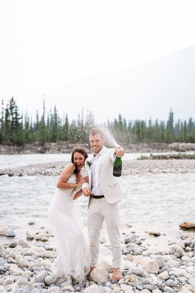 Groom sprays champagne at this Jasper Intimate Wedding Photographer as they stand next to the Snaring River. 