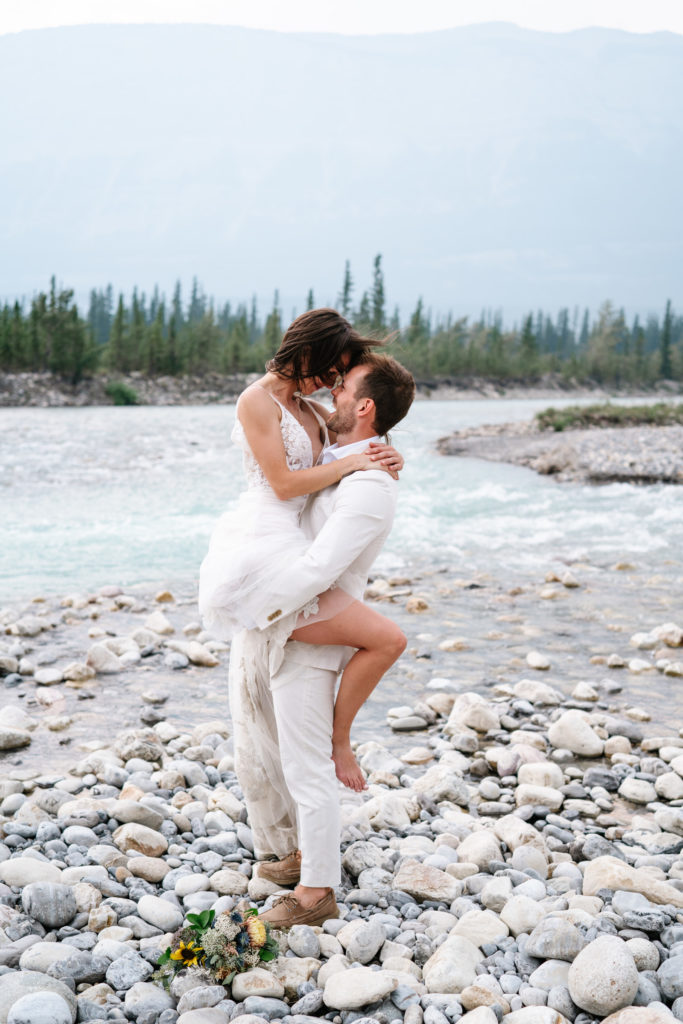 groom in white suit holds bride in lace wedding up next to the Snaring River in Jasper National Park