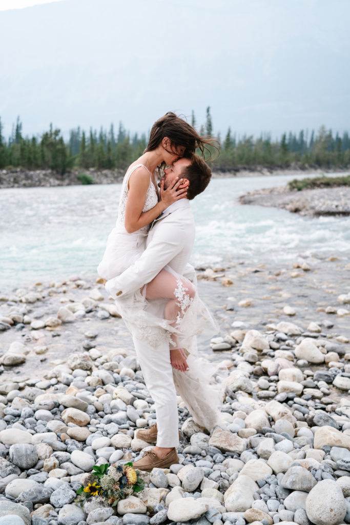 bride kisses grooms head as he holds her up barefoot in Jasper National Park