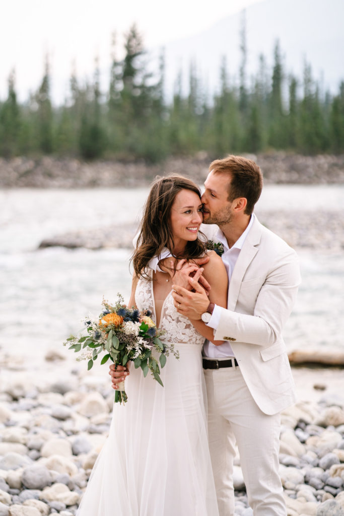 groom embraces bride from behind and kisses her temple while she looks off into the distance in Jasper National Park while holding her flowers. 