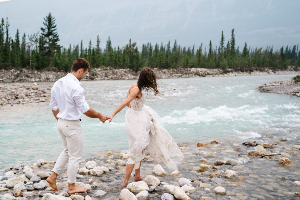 Barefoot bride leads barefoot groom along rocks next to Snaring river in Jasper National Park.