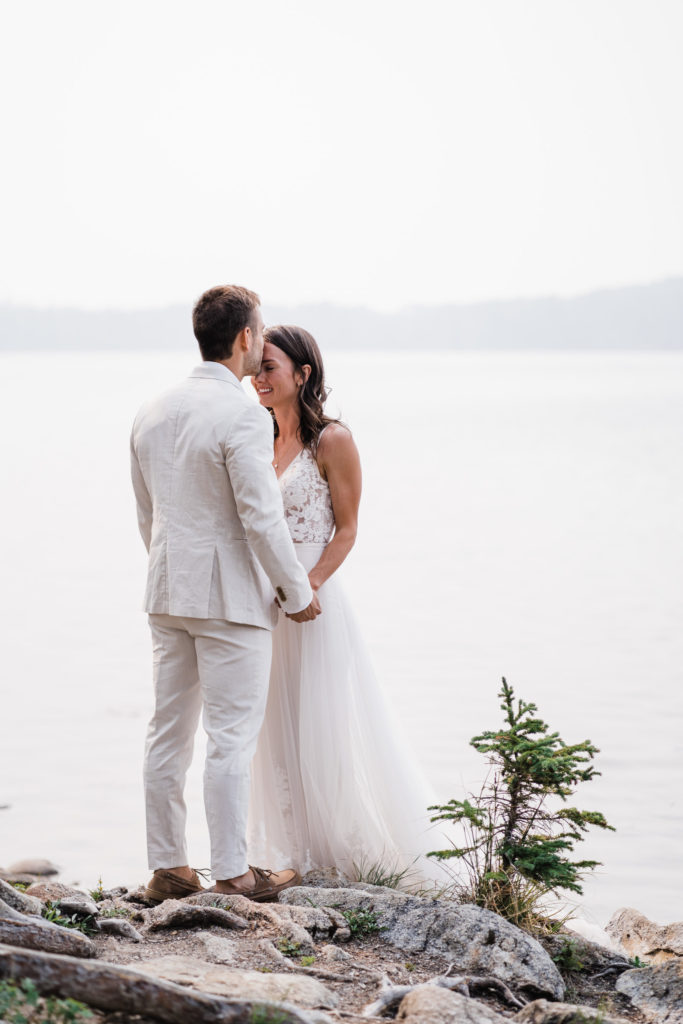 Groom kissing brides forehead while waterfront at Pyramid Island in Jasper National Park.