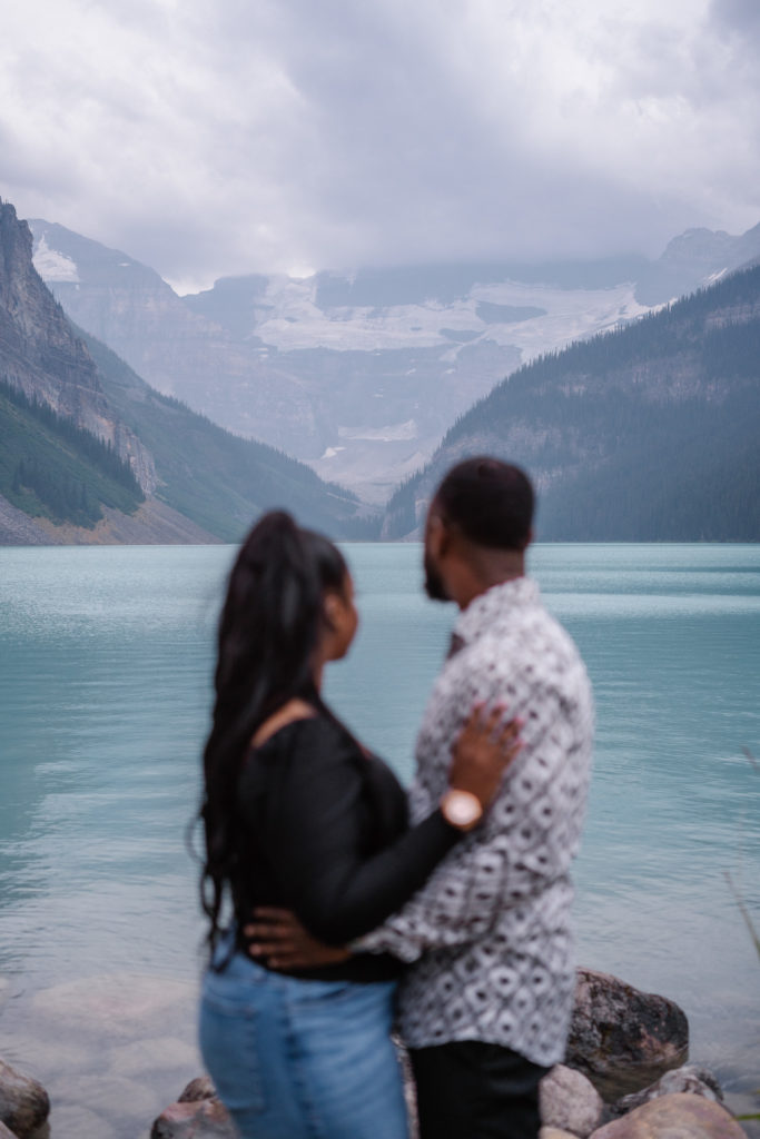 Couple hug and look out at Lake Louise with the focus on the glacier in the back. 