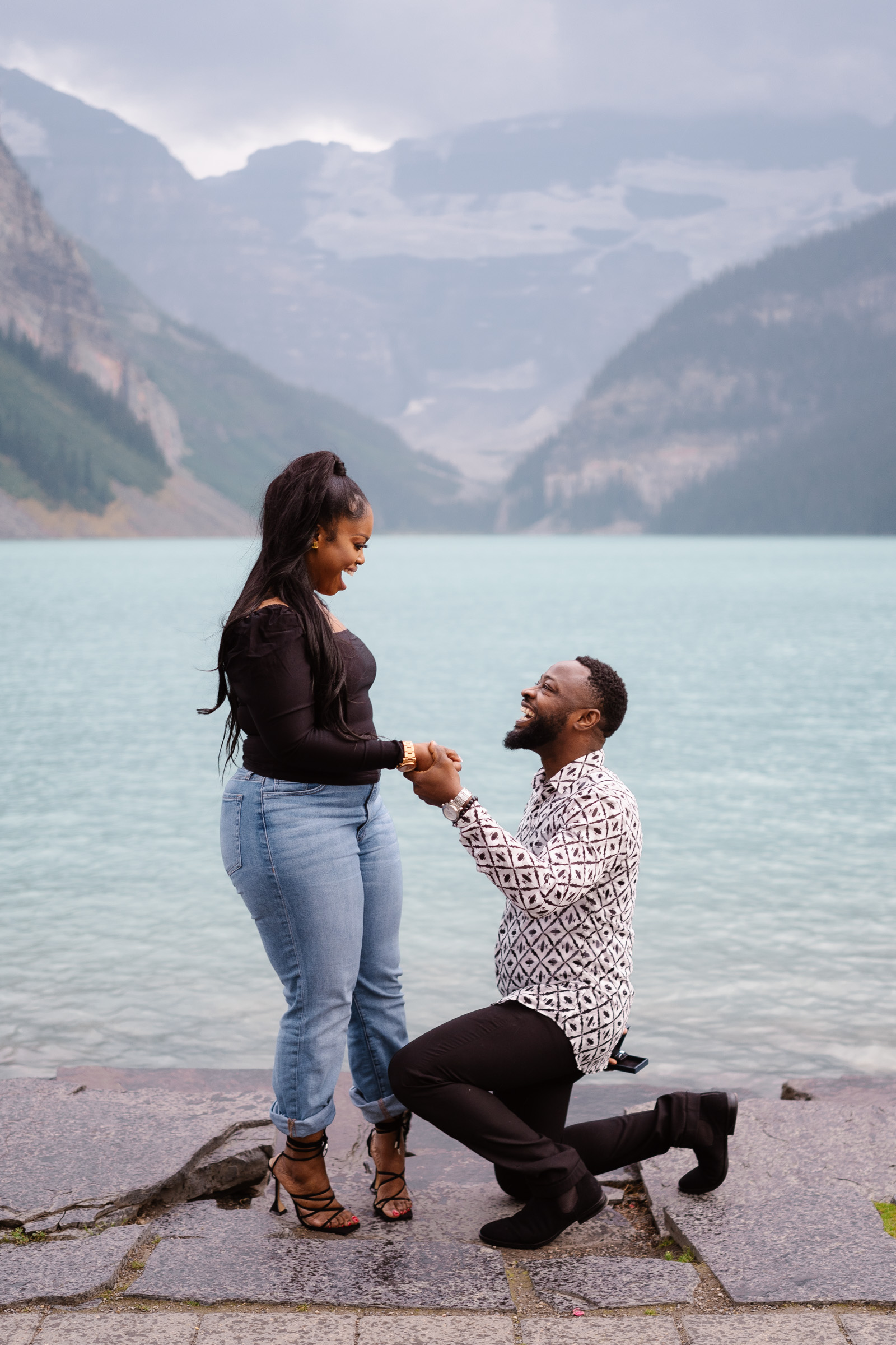 Man is down on one knee infront of Lake Louise while woman holds his hand with a surprised look on her face. 