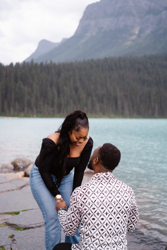 Woman bends over in disbelief as boyfriend proposes at Lake Louise in Banff National Park.