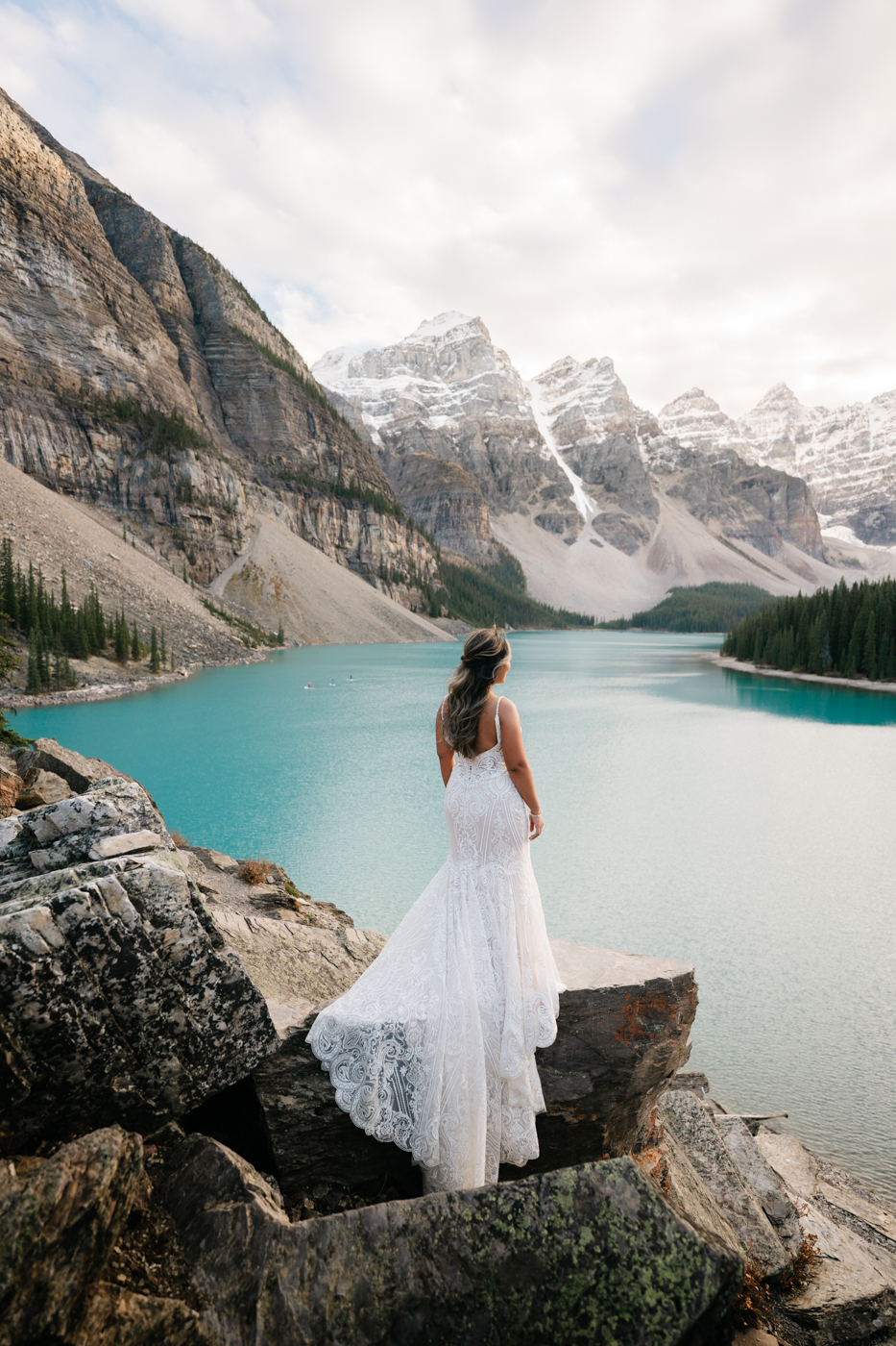 Brides lace dress trails behind her on rocks at Moraine Lake