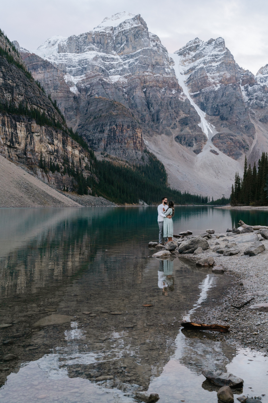 Couple stands on shore of Moraine Lake with a reflection of them and the mountains. 