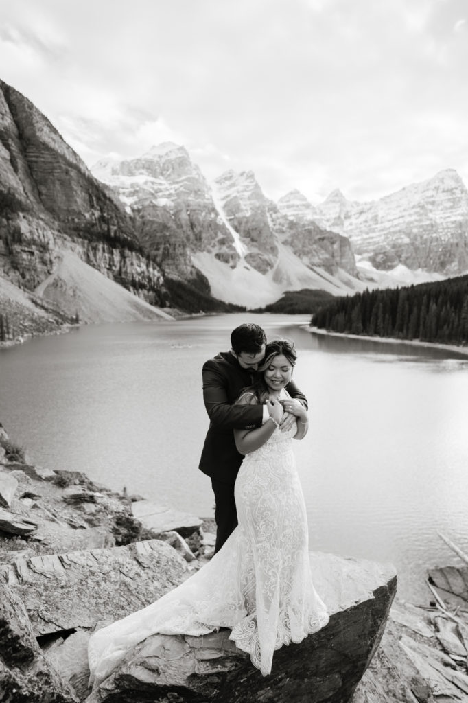 Black and white image of groom cuddling behind bride while on rocks at Moraine Lake