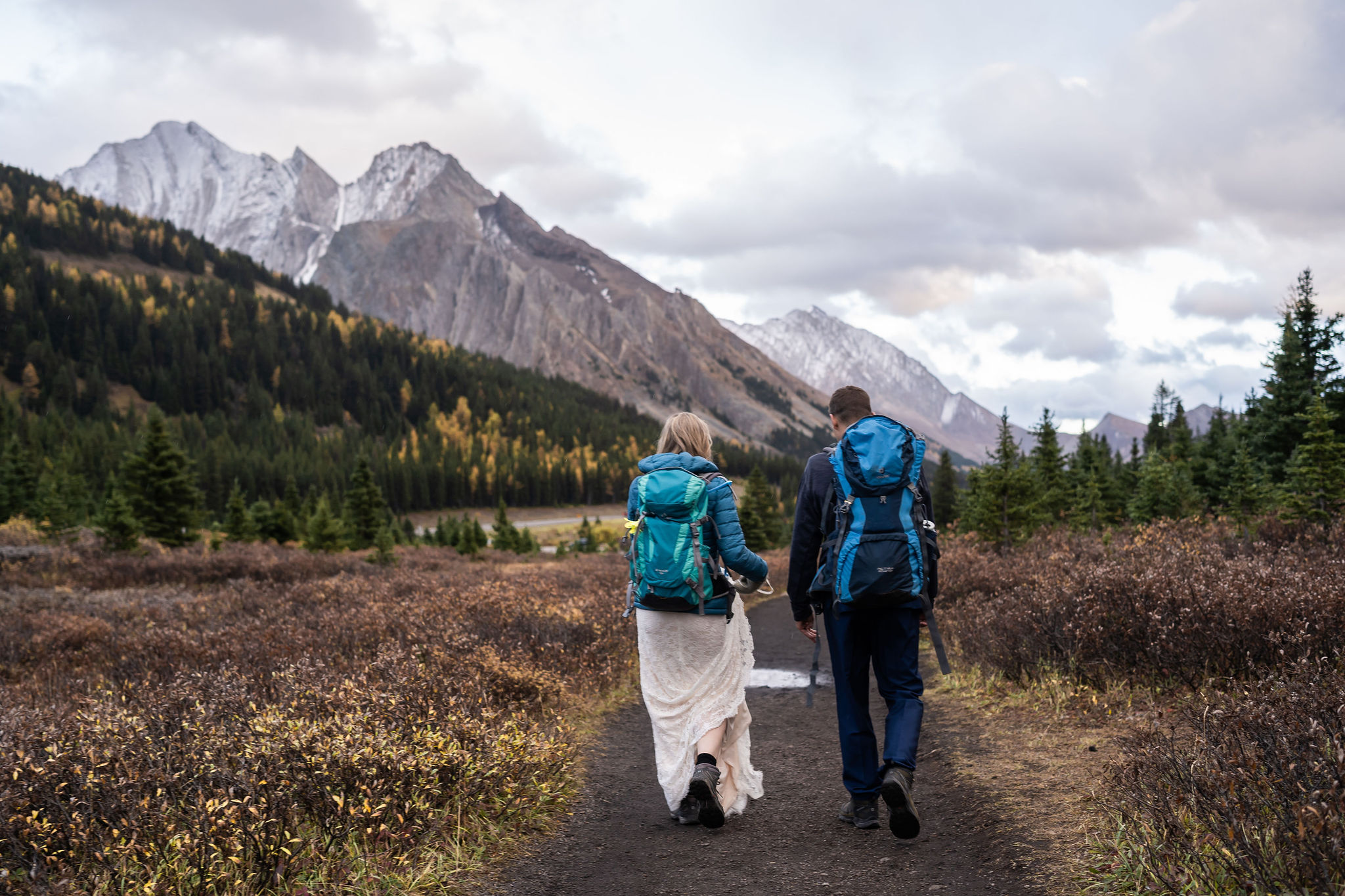 Bride and groom wear backpacks walking down trail after their kananaskis hiking elopement