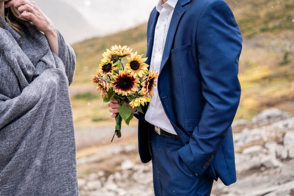 groom in blue suit holds bouquet of sunflowers.