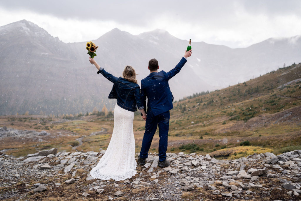 Bride and groom with arms up look down the valley as the snow falls in Kananaskis 