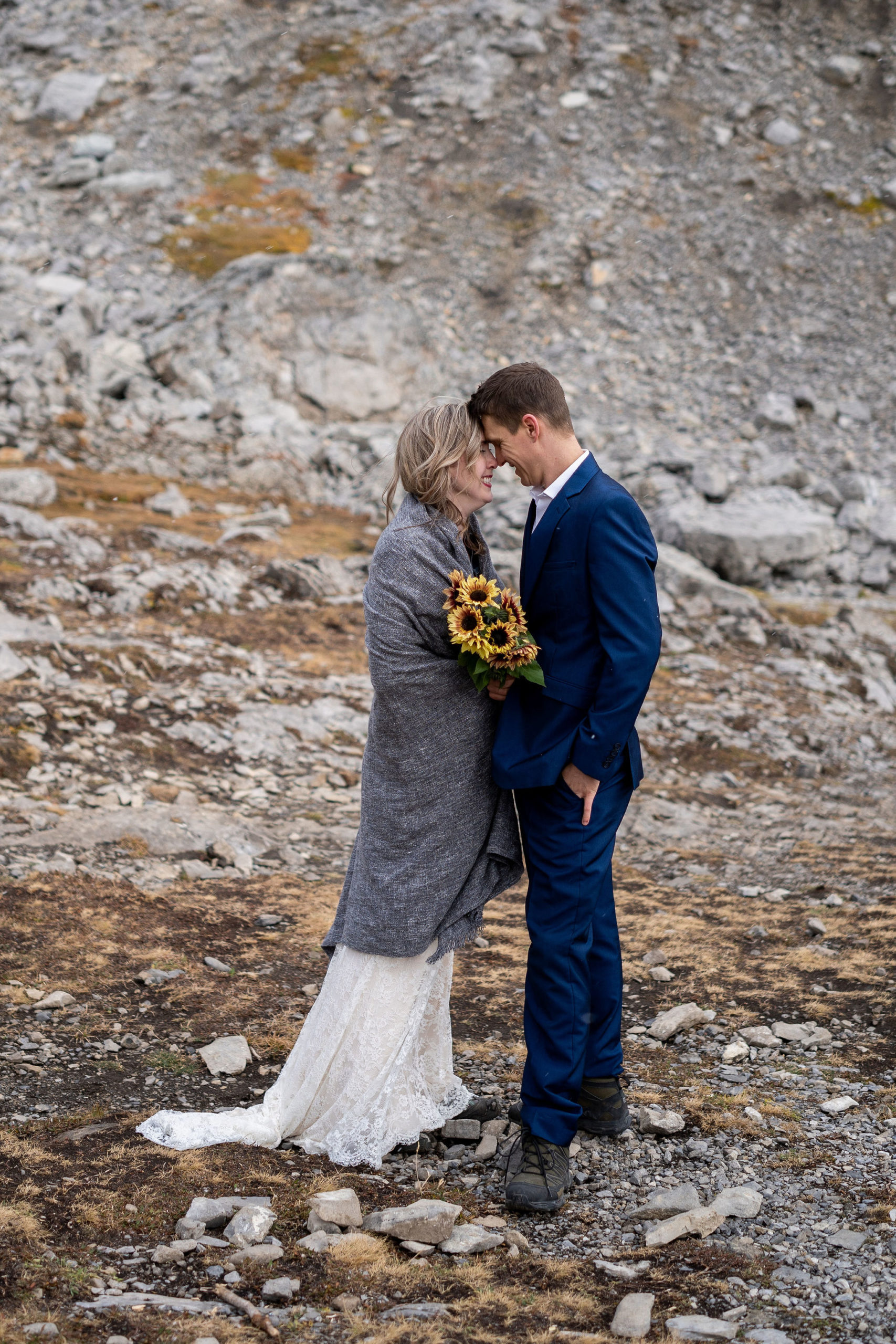 Bride wrapped in grey blanket holding sunflowers touches her forehead to the groom as they smile