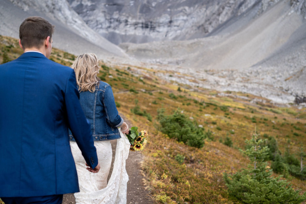 Groom and bride walk along Ptarmigan Cirque in the fall as she carries a bouquet of sunflowers. 
