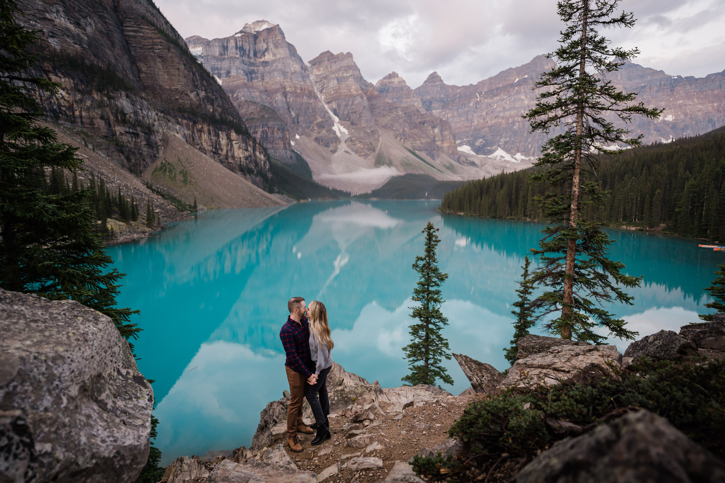 Couple holds hands facing each other and kissing in front of Moraine lake before sunrise. 